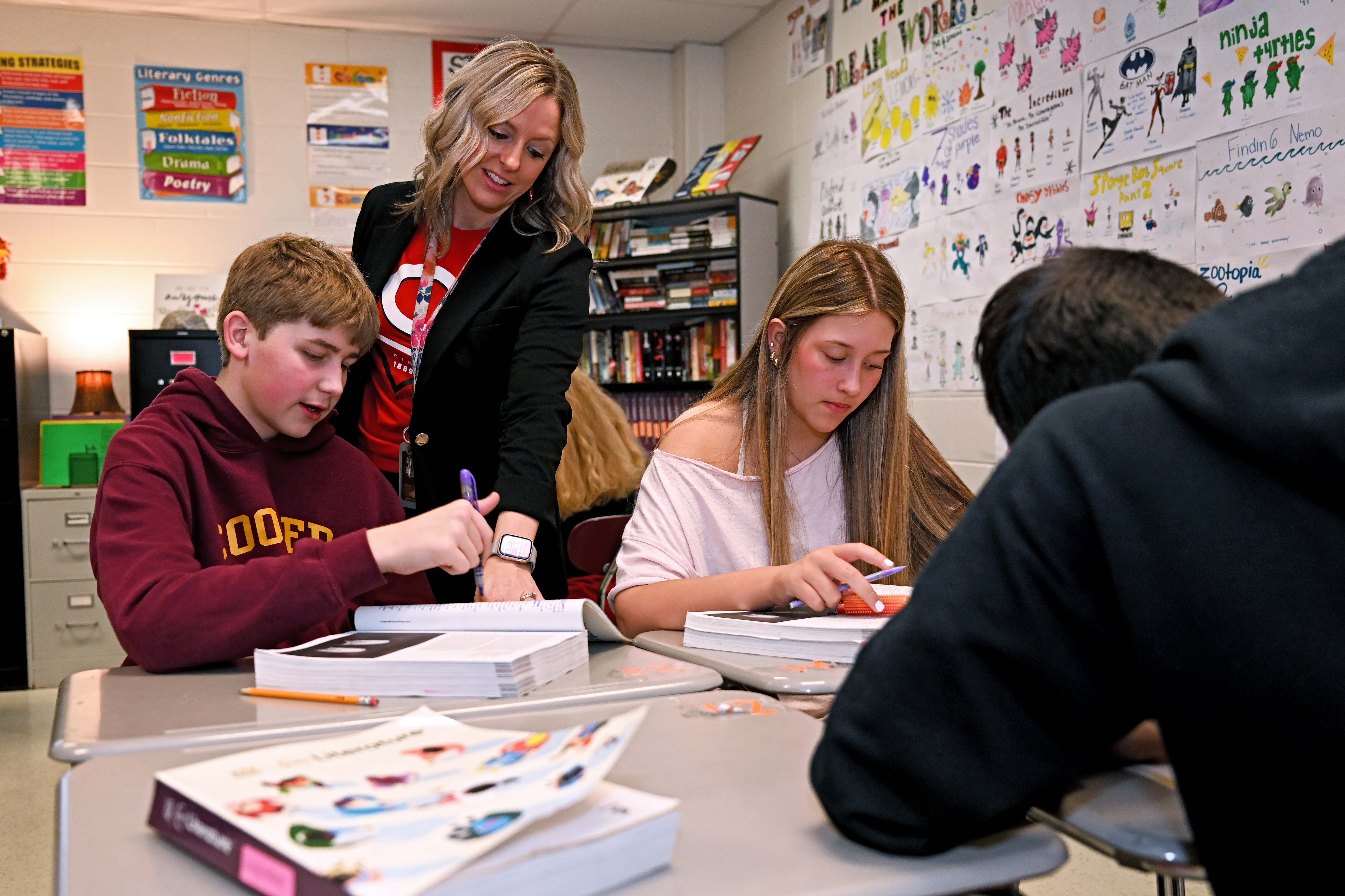 A teacher stands beside students working at desks in a classroom, leaning in to support them as they complete an assignment. Books, student work and colorful instructional materials are visible throughout the room.